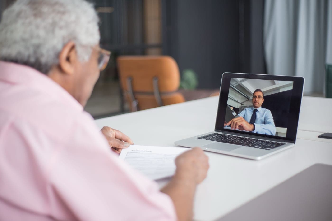 A man at his desk on a video call, a printed form in his hand ready to sign